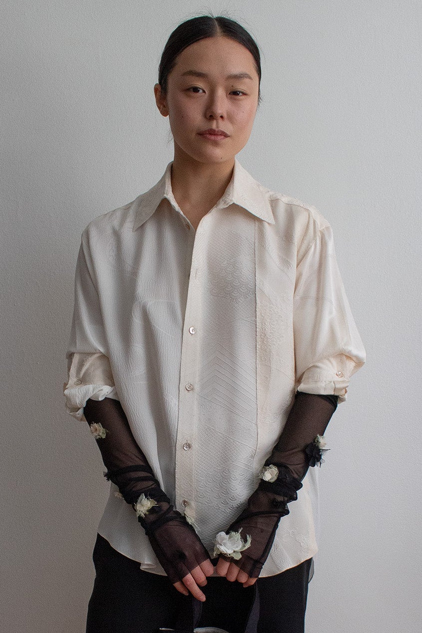 Person wearing a white shirt and black skirt with floral accessories against a plain wall.