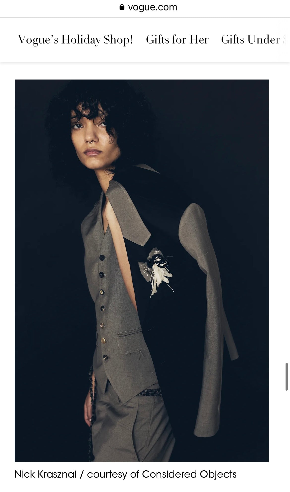 A fashion editorial photo from Vogue.com showing a model wearing a Considered Objects dark gray tailored suit with a structured vest and matching trousers against a very dark background.