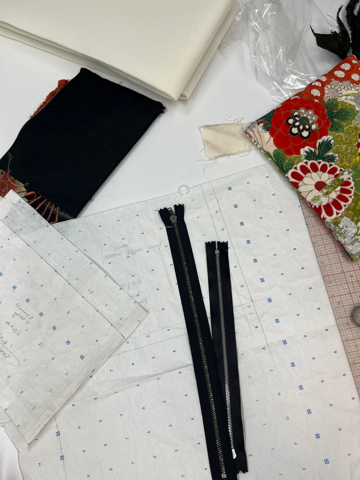 Two black metal zipper pulls laid on top of patterned fabric, surrounded by sewing materials including folded cream fabric, a black fabric swatch, a floral fabric swatch, and a piece of gridded paper on a white surface.