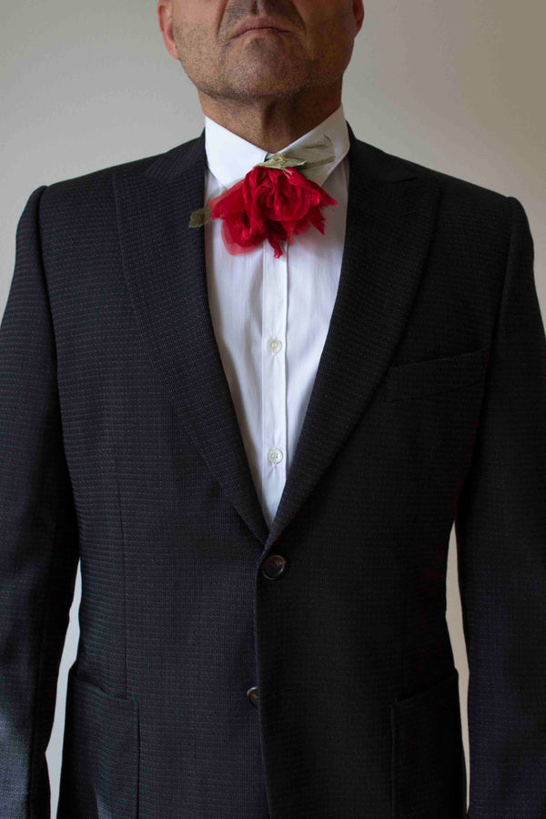 Man wearing a dark suit with a white shirt and red floral bow tie against a plain background