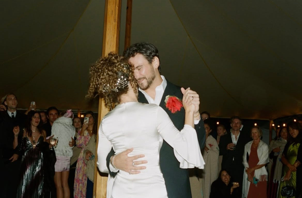 A couple dancing during a wedding reception inside a tent, with guests visible in the background. The man is wearing the Falling Flower brooch in ume red. 