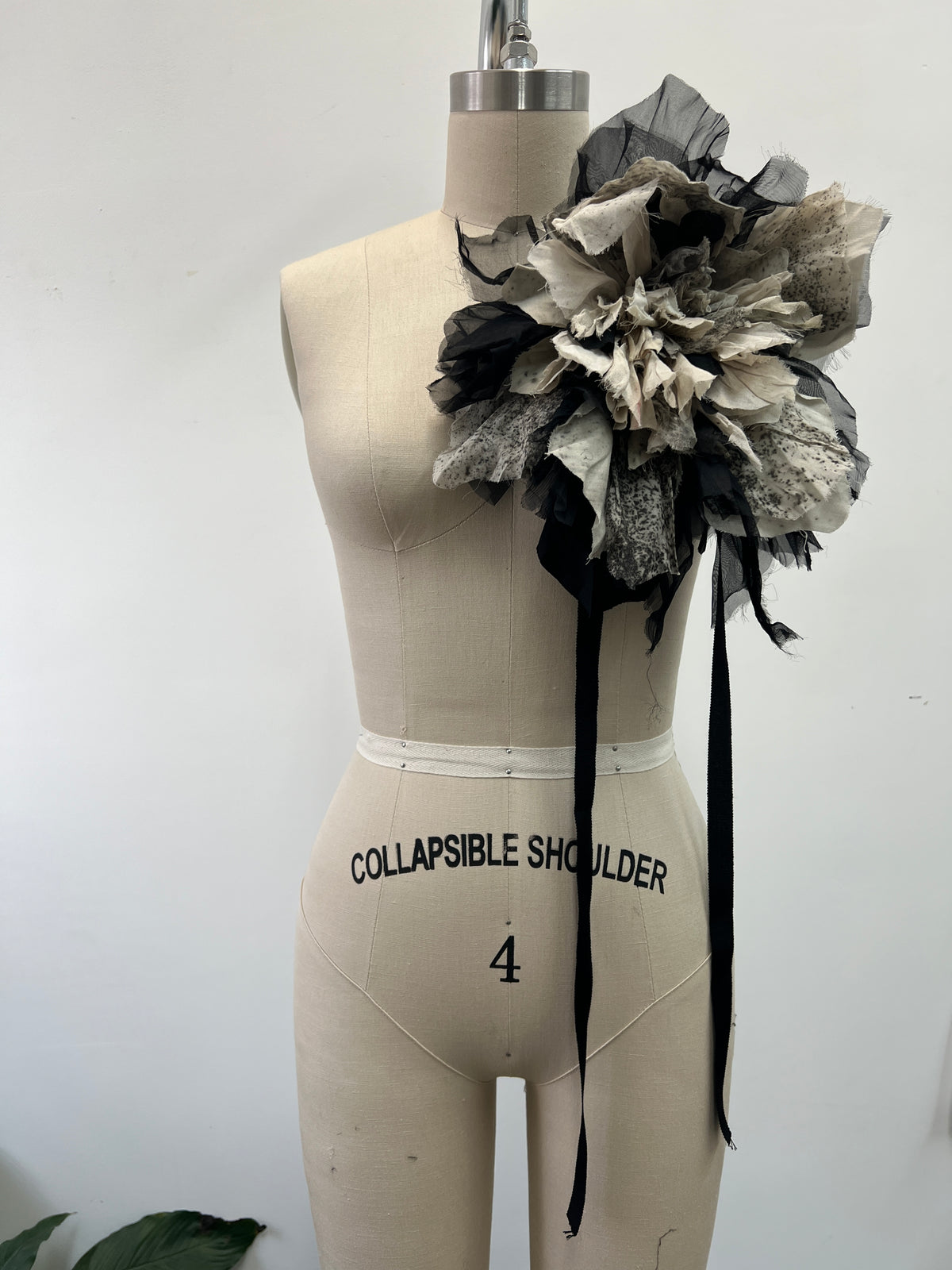 A large sculptural shoulder flower embellishment made of layered black, gray, and off-white tulle and fabric petals, attached to a beige dress form mannequin. The dress form has 'COLLAPSIBLE SHOULDER 4' printed on the front.