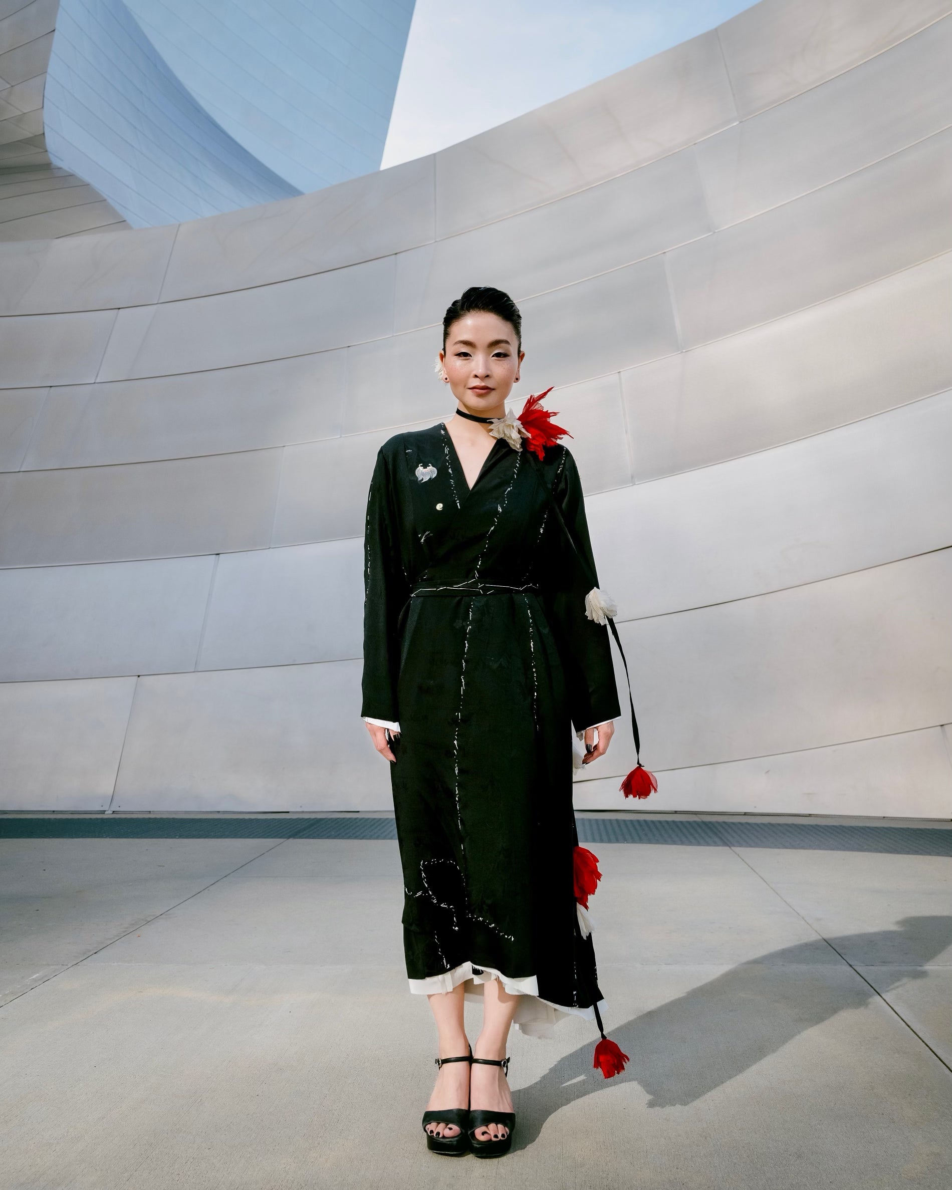 A woman stands outdoors in front of a curved modern metallic wall, wearing the Irregular Hand-stitched Kimono Wrap Dress, Falling Flowers necklace in ume red/ivory.  Photography by Alex Shibutani, courtesy of Maia Shibutani. 