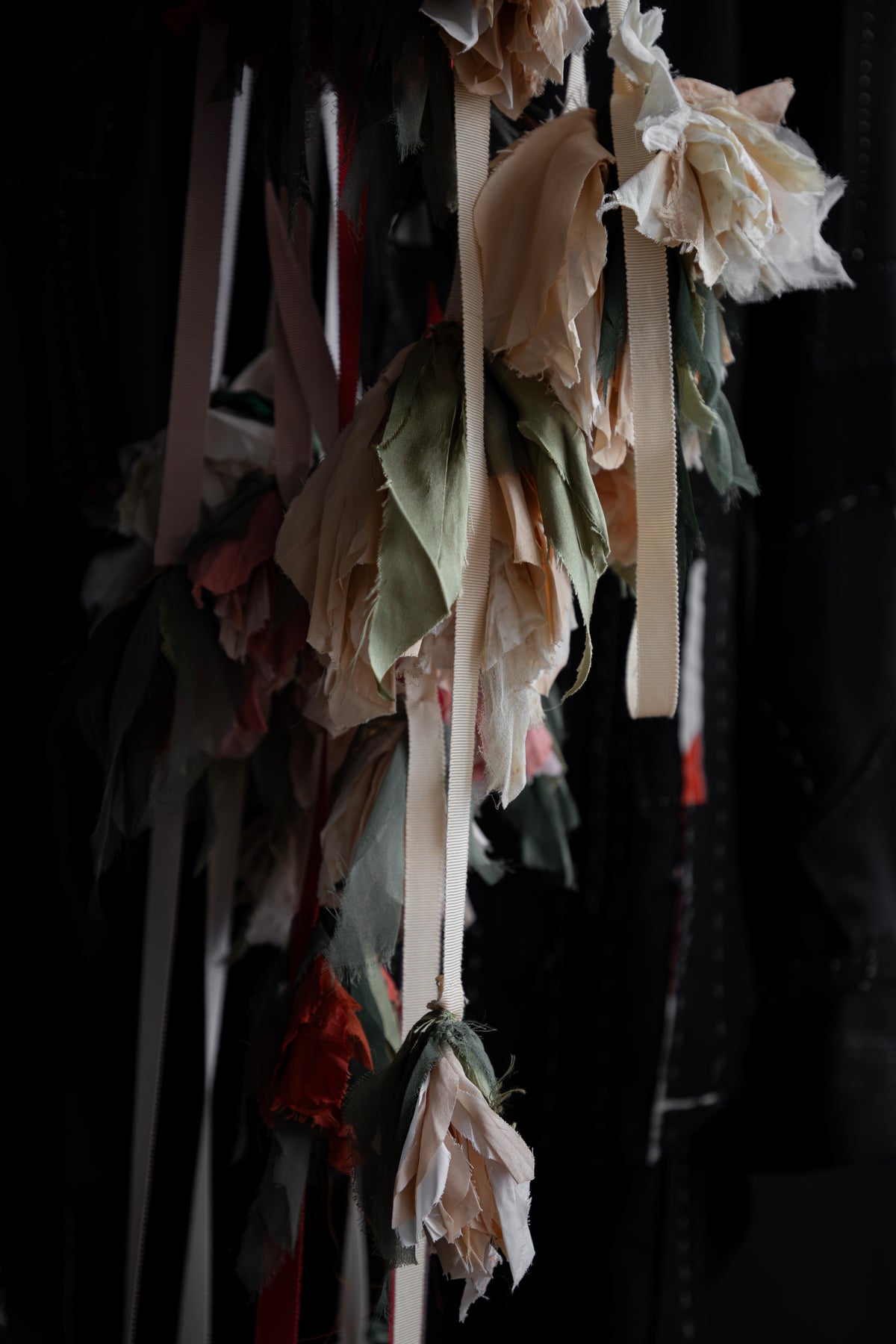 A close-up of hanging fabric flower garlands made from layered, tattered off-white and pale pink petals with muted green and red fabric leaves, set against a very dark background.