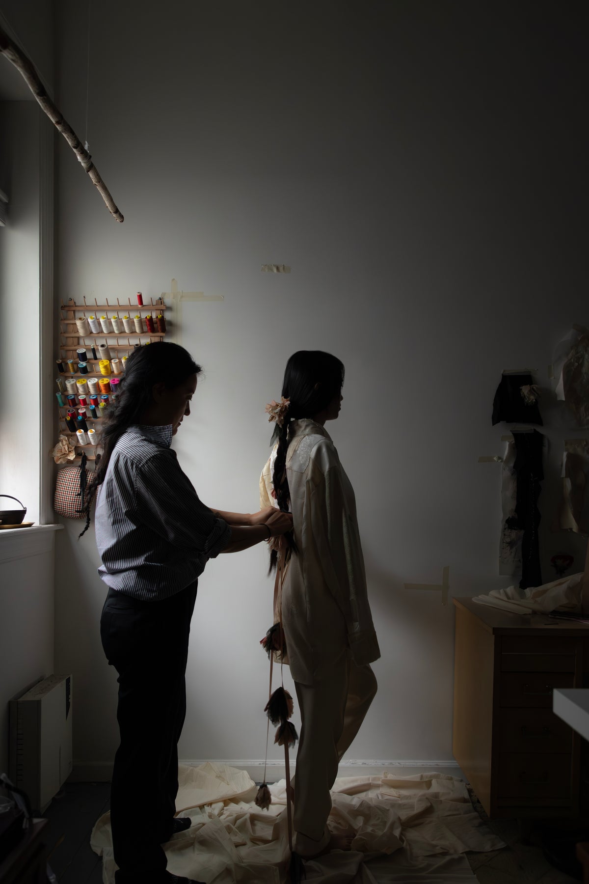 A person in a long cream-colored outfit stands in profile while another person adjusts their long dark hair in a dimly lit studio room with shelves and sewing materials visible in the background.  Considered Objects studio. 
