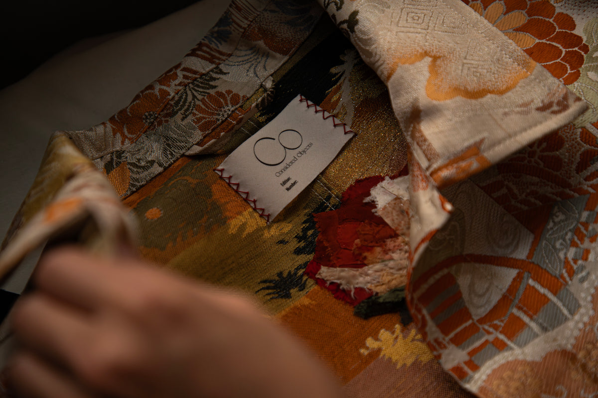 A close-up of a patterned textile with a visible sewn-in label reading Considered Objects, showing a hand holding part of the fabric. The obi material has an abstract floral and geometric print in orange, beige, brown, black, and muted red tones.