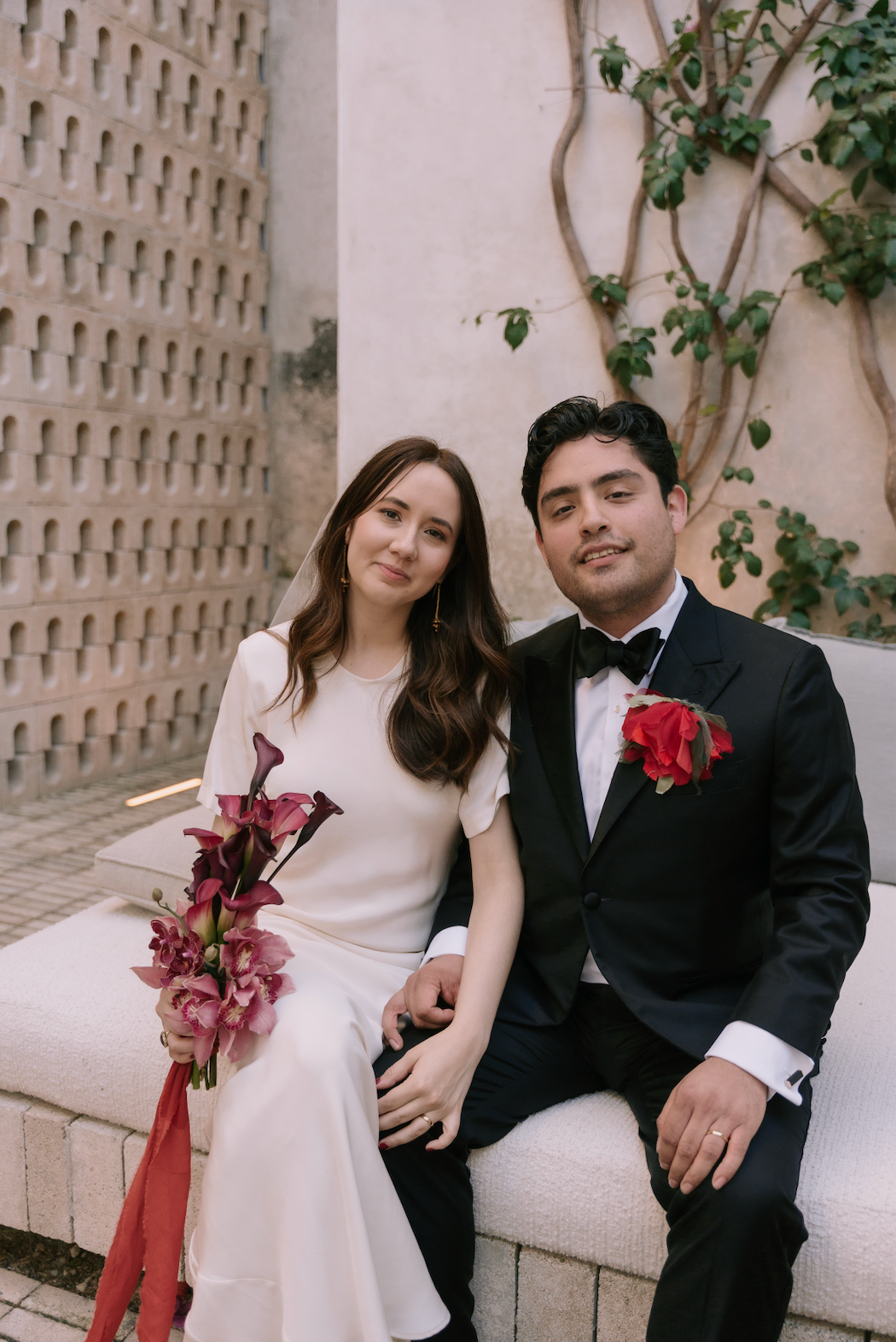 A bride and groom are sitting together on a stone bench. The groom is wearing the Falling Flower brooch in ume red.  Photography by Gaby Gonzales, courtesy of Monica Biles.